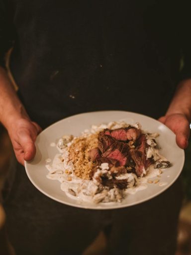 A person holding a plate of beef dish with blurred festive lights in the background.