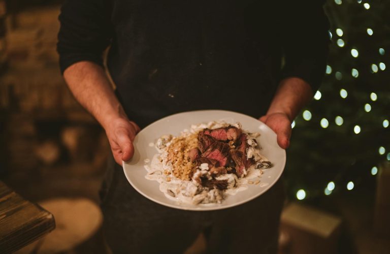 A person holding a plate of beef dish with blurred festive lights in the background.